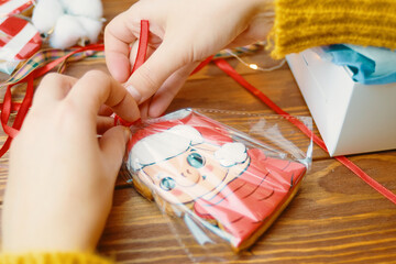 Close up of cookies with colored icing on wooden table. Christmas gifts. Women's hands tie red ribbon on bag of gingerbread in form of pig in Santa Claus hat. New Year's atmosphere.