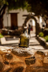 Vertical format photograph of a bottle of green wine without content on a small white wall in a square in the town of Cachi, Salta, Argentina.
