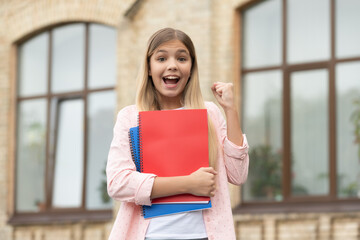 Happy teen girl with school books making winning gesture outdoors, victory