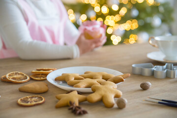 Fototapeta na wymiar Christmas traditional cookies stars, hearts, men on a white plate. On the wooden table are dry oranges, anise, cookie molds and tassels. Children's hands, Christmas tree with lights in the background