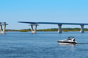 A small speedboat floats along the river against the backdrop of a bridge under construction. Sunny...