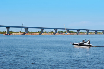 Fototapeta premium A small speedboat floats along the river against the backdrop of a bridge under construction.
