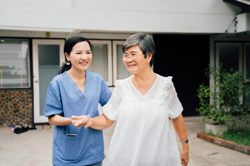 Optimistic female nurse in blue uniform supporting Asian elderly woman while walking together through gate outside house