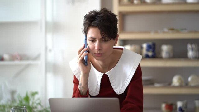 Italian woman sculptor working at laptop, checking orders at her online shop, talking with client at smartphone in comfortable workplace. Freelancer female take peaceful break for relaxation