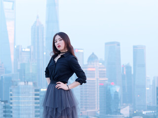 Beautiful young Chinese woman in black posing on top of mansion roof with blur Shanghai Bund landmark buildings background with dusk light. Emotions, people, beauty and lifestyle concept.