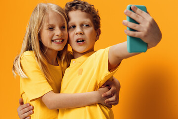 cute school children brother and sister take funny selfies on their smartphone while standing in bright clothes on an orange background
