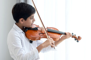 Indian boy practicing playing violin at home, holds the violin instrument beside the window,...