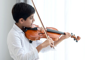 Indian boy practicing playing violin at home, holds the violin instrument beside the window, creative children play song, classical education