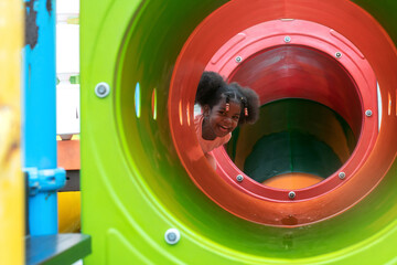 Happy African child girl playing in a tunnel at outdoor playground in the park on summer vacation,...