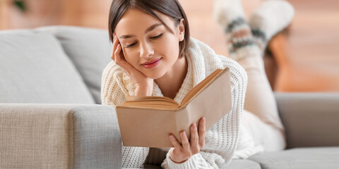 Beautiful young woman reading book while lying on sofa at home
