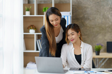 Two professional Asian business women discuss work using a laptop computer to work in the office.