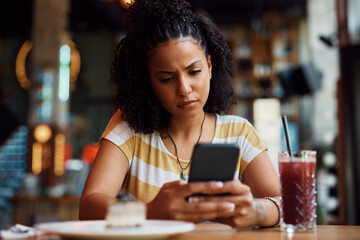 Distraught African American woman reading text message on mobile phone in cafe.