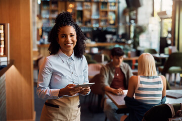 Happy black waitress using digital tablet while working in cafe and looking at camera.