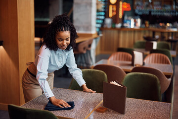Happy African American waitress cleaning tables in cafe.