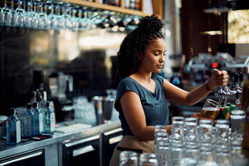 Young black waitress pours beer draft beer while working at bar counter.