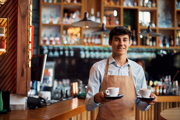 Young waiter serving coffee in cafe and looking at camera.