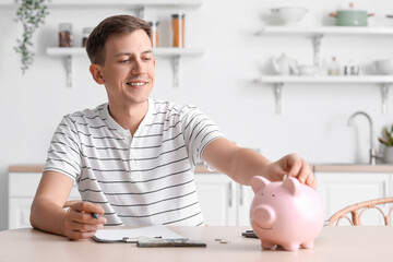 Young man putting coin into piggy bank at table in kitchen
