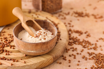 Board with bowl of flour, scoop and buckwheat grains on beige background, closeup