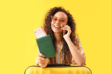 Young African-American woman with passport and suitcase talking by mobile phone on yellow background