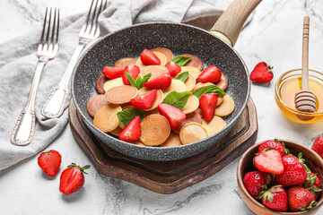 Wooden board with frying pan of mini pancakes and strawberry on light background