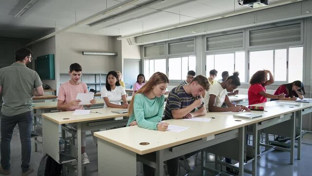Young teacher giving test sheets to students in class. Multi ethnic teenager pupils ready to take exam in college high school classroom
