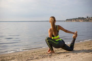 Rear view of girl doing gymnastics on the beach. Blonde doing yoga on the river bank. Young woman does leg stretching in sport outfit