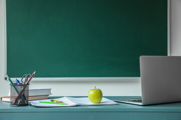 Apple with books, pen cup and laptop on table near school chalkboard
