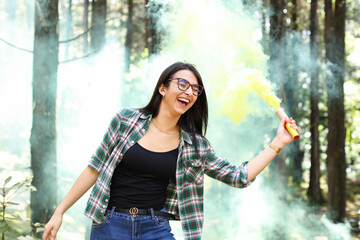 Young woman in a photoshoot raising her hand holding a smoke bomb yellow in the forest smiling at...