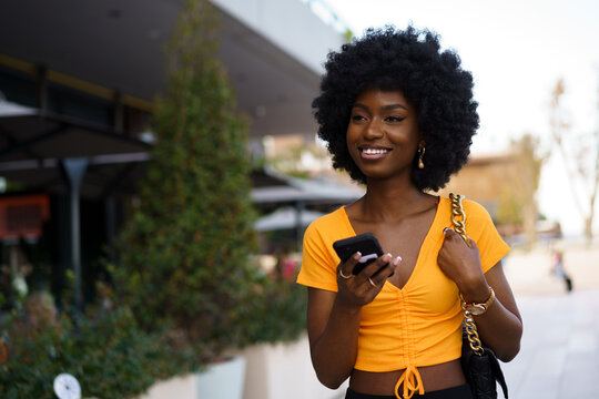 Smiling afro-american woman holding mobile phone while walking in the street