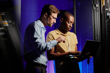 Waist up portrait of two network engineers using computer in dark server room lit by neon light,...