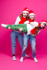 Seasonal Holidays Ideas. Three Happy Caucasian Girls Wearing Santa Hats Having Fun While Having Teenare Girl Lifted On Hands With Wrapped Golden Gift.
