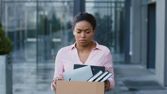 Dismissal concept. Young stressed fired african american manager walking from office, carrying box with personal stuff