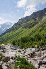 Fototapeta premium A small wooden bridge across the stormy river Terskol and rocky banks among the slopes of mountains and green grass on a bright sunny summer day near Elbrus region