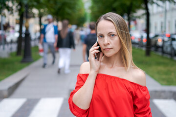 Attractive businesswoman talking on a mobile phone with a client standing on a city street during the day in a red dress. Caucasian woman succeeding