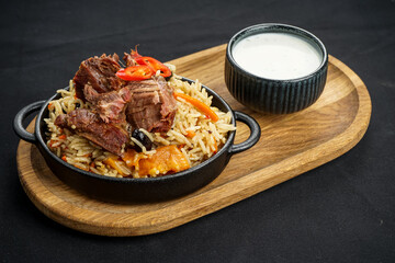 rice pilaf with meat in a round pan on a board with sour cream on a black background
