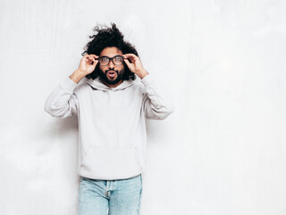 Handsome smiling hipster  model.Sexy unshaven Arabian man dressed in summer stylish hoodie clothes. Fashion male with long curly hairstyle posing near grey wall in studio. In spectacles, glasses