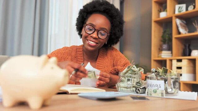 Finance, budget and saving money by depositing cash into a piggy bank after calculating a budget in a home living room. Smiling, happy and cheerful woman with afro planning her spending and expenses