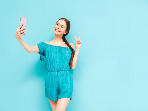 Young beautiful smiling female in trendy summer blue overalls. Sexy carefree woman posing near wall in studio. Positive brunette model holding smartphone. Taking selfie photos