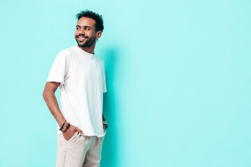 Fototapeta premium Handsome smiling hipster model. Sexy unshaven man dressed in white summer t-shirt and jeans clothes. Fashion male posing near blue wall in studio. Isolated