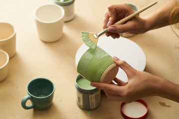 Close-up of girl painting clay mug with glaze. Woman coloring pottery in workshop with a...