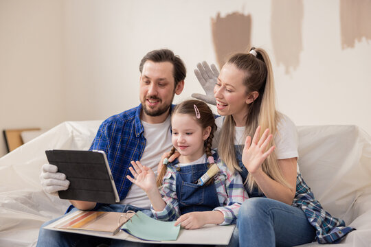 Happy family dressed in denim style sitting on a sofa in a room undergoing renovation. They went online to communicate with their friends, whom they wave so happily.