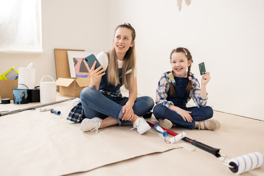 Two girls are sitting on floor in room being renovated. One blonde girl, 27 years old, is holding a phone and thinking about something. The second girl is 8 years old and is holding a credit card.