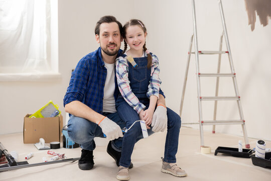 Little girl 8 years old wearing denim overalls and checkered shirt sits on dad's lap. Handsome middle-aged man smiling and holding paint roller in hand. In background are various things for repair.