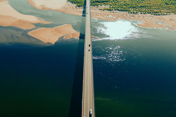 Aerial drone shot of cars driving on bridge over river and sandy riverside at sunset. Amazing aesthetic 4k top view shot with beautiful coast with dune sands. Drone aerial bridge with car traffic