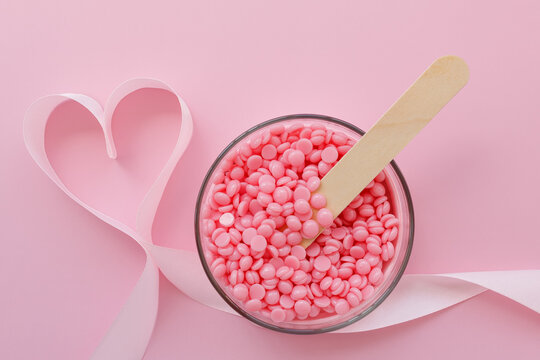 Beautiful granules of pink wax for depilation in a glass bowl, a wooden spatula and a ribbon in the shape of a heart on a pink background. I love waxing. Epilation, depilation, unwanted hair removal.