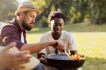 A bearded middle-aged guy is preparing a barbecue for friends. A group of college guys spend time together surrounded by nature.