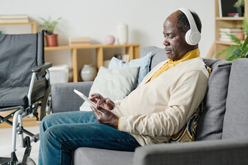 African man with disability sitting on sofa in wireless headphones and using tablet pc for listening music in the room