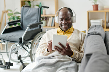 African senior man in wireless headphones typing on digital tablet and smiling while lying on sofa in living room