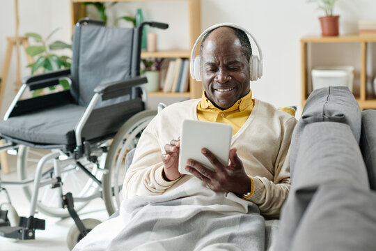 African senior man in wireless headphones typing on digital tablet and smiling while lying on sofa in living room