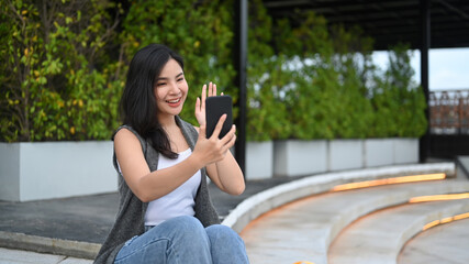 Cheerful asian woman having video call with friend on mobile phone while sitting in a park on a sunny day